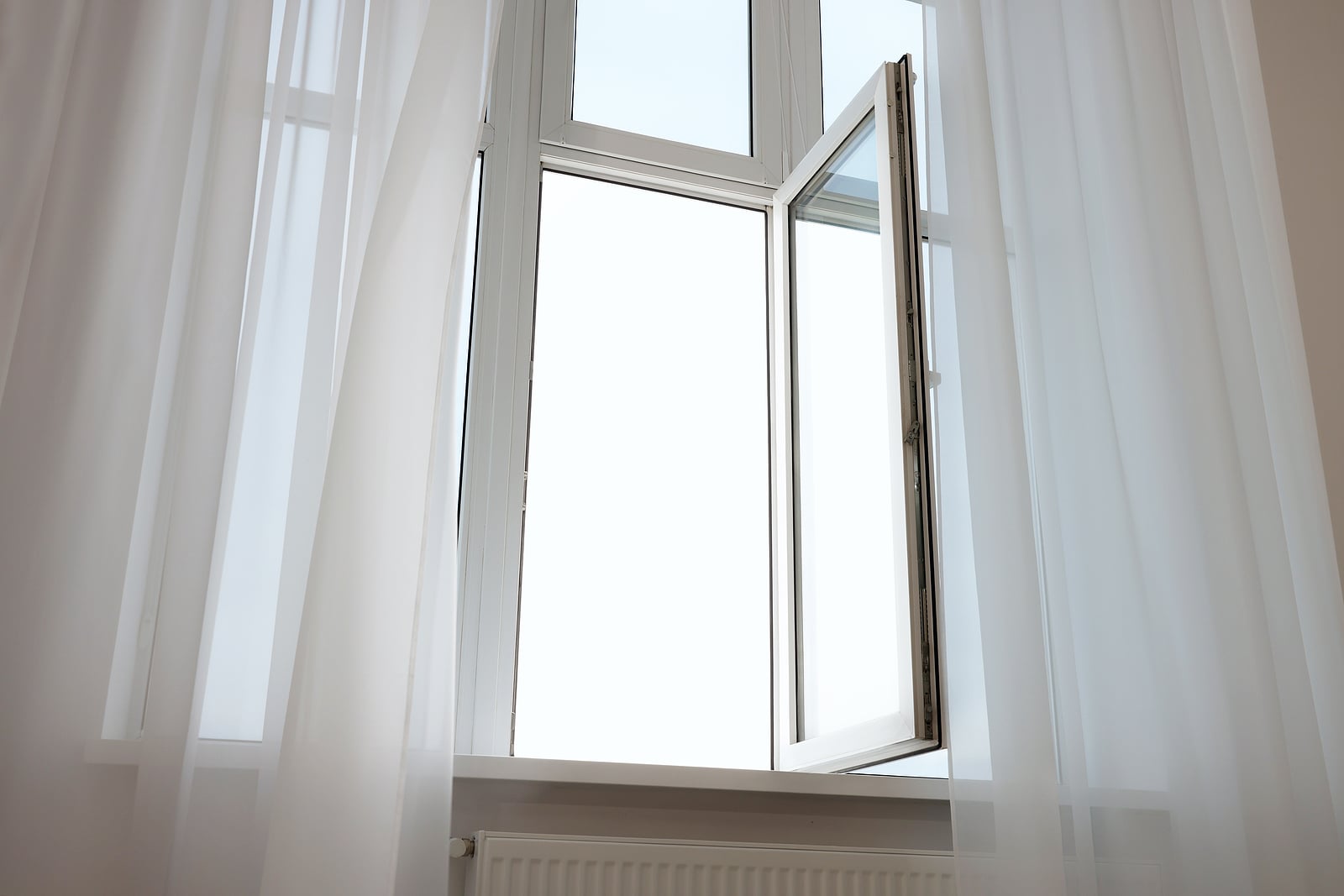 An open white-framed inswing window bordered by sheer white curtains 