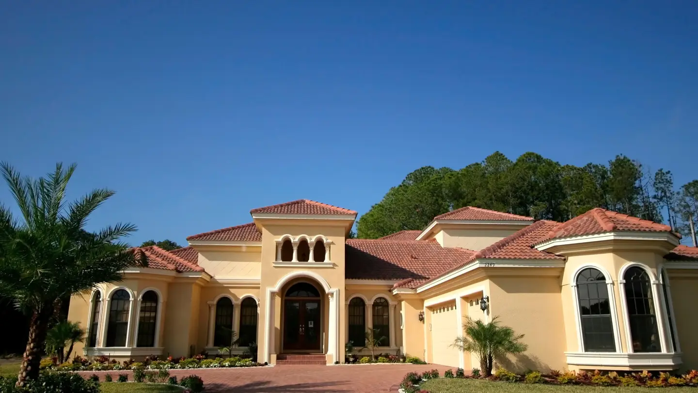 Large brown Florida home with large windows and bushes, palm tree, and grass in front.